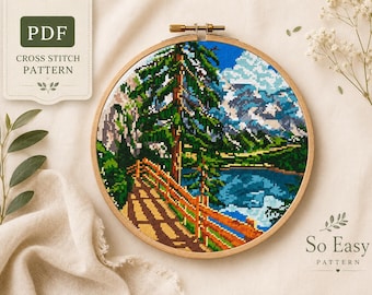 May include: A cross-stitch pattern depicting a mountain lake landscape, held in a wooden embroidery hoop. The design shows a bridge, trees, and mountains, with a blue sky and water. The text "PDF CROSS STITCH PATTERN" is visible.