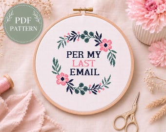 Könnte beinhalten: Ein runder Stickrahmen aus Holz mit weißem Stoff und einem Kreuzstichmotiv. Das Design zeigt einen Blumenkranz in Dunkelblau, Grün und Rosa, der den schwarzen und rosafarbenen Text PER MY LAST EMAIL umschließt.