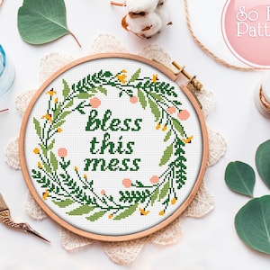 May include: A white embroidery hoop with a green floral wreath and the text "Bless this mess" in green lettering. The hoop is on a white surface with a piece of lace and green leaves.