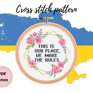 Könnte beinhalten: Kreuzstichmuster mit einem Blumenkranz mit dem Text "This is our place, we make the rules" in einem goldenen Rahmen. Der Kranz befindet sich auf einem blauen und gelben Hintergrund, der der ukrainischen Flagge ähnelt.