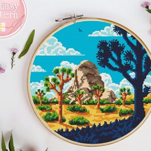 May include: Cross stitch pattern of a desert landscape with a large rock formation, Joshua trees, and a blue sky with clouds. The pattern is framed in a wooden hoop.