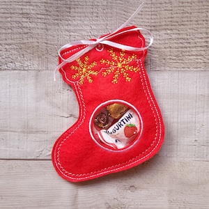 May include: Red felt Christmas stocking with a clear plastic window, decorated with yellow snowflakes and filled with candy.