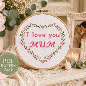 Peut inclure: Un cercle de broderie au point de croix avec les mots "I love you Mum" en rose, entourés d'un cœur floral. Le cercle est présenté sur une surface en dentelle, avec un vase de fleurs roses et des photos encadrées en arrière-plan.