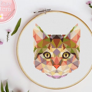 May include: A geometric cross-stitch pattern of a cat's face, featuring a mosaic of colorful triangles in shades of orange, green, purple, and brown. The cat has yellow eyes and is framed in a wooden embroidery hoop. The text "So Easy Pattern" is in the upper left corner.