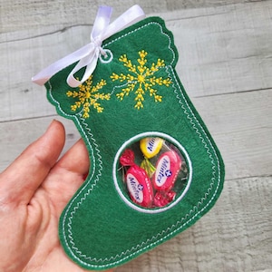 Può includere: Calza di Natale in feltro verde con una finestra di plastica trasparente che mostra carameli. La calza ha cuciture bianche e fiocchi di neve ricamati in giallo.