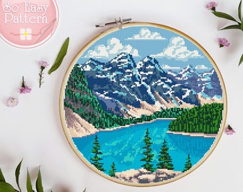 May include: Cross-stitch pattern of a mountain lake scene with blue water, green trees, and snow-capped mountains. The pattern is framed in a wooden hoop.