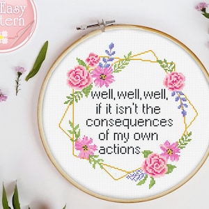 May include: A white embroidery hoop with a gold geometric frame and pink and purple flowers. The text inside the frame reads "well, well, well, if it isn't the consequences of my own actions."