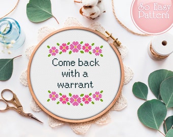 May include: A white embroidery hoop with a cross-stitch design that reads "Come back with a warrant". The design is surrounded by a border of pink flowers.