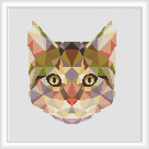 May include: A framed cross-stitch of a cat's face, featuring a geometric design. The cat's face is composed of various colored triangles, including shades of green, brown, purple, and yellow. The cat has large, round eyes and a small pink nose. The frame is white.
