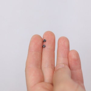 Könnte beinhalten: Nahaufnahme von zwei kleinen, dunkelgrauen Ohrringen, die zwischen zwei Fingern vor weißem Hintergrund gehalten werden. Die Ohrringe haben ein kreisförmiges Design mit einem kleinen, zentralen Detail. Das Bild zeigt die Details des Schmucks.