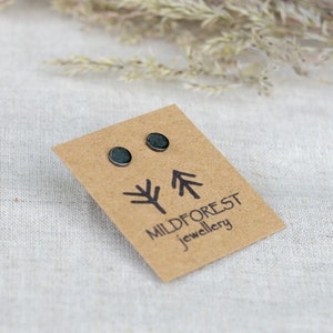 Può includere: Un paio di piccoli orecchini rotondi di colore verde scuro su una carta marrone con il testo "Mildforest jewellery" e un design stilizzato di un albero.