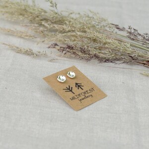 Puede incluir: Un par de pendientes de bot&oacute;n de plata en una tarjeta marr&oacute;n con el texto "Mildforest Jewellery" y un dise&ntilde;o de &aacute;rbol estilizado.