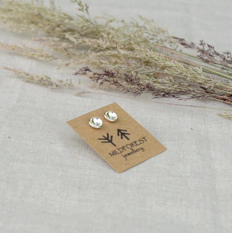 Puede incluir: Un par de pendientes de bot&oacute;n de plata en una tarjeta marr&oacute;n con el texto "Mildforest Jewellery" y un dise&ntilde;o de &aacute;rbol estilizado.