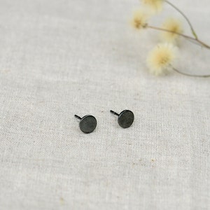 May include: A pair of black stud earrings with a circular design. The earrings are on a white fabric background.