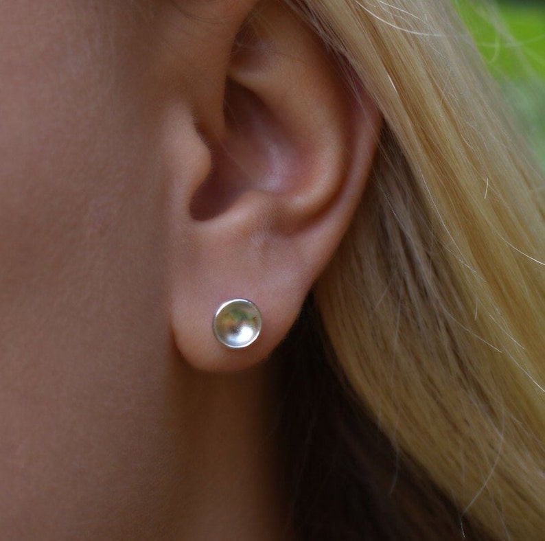Puede incluir: Un par de pendientes de plata con un dise&ntilde;o redondo ligeramente c&oacute;ncavo. Los pendientes se llevan en la oreja de una persona.