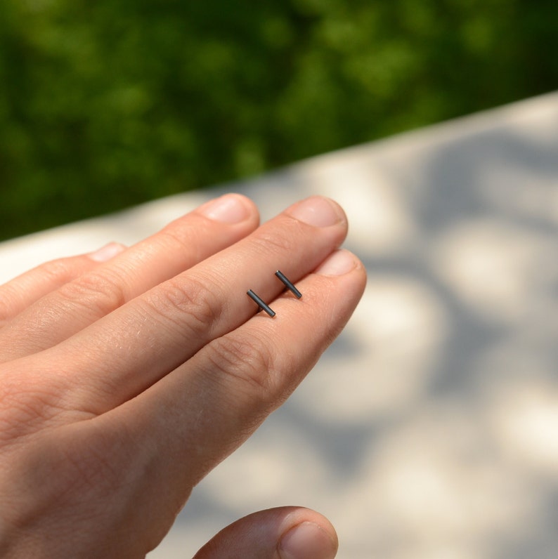 May include: A pair of small, black bar stud earrings. The earrings are shown on a person's hand.