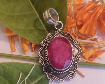 May include: A silver pendant with a red gemstone set in a decorative frame. The pendant is surrounded by orange flower petals.