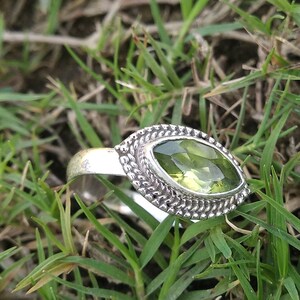 Op de afbeelding: Een zilveren ring met een groene edelsteen gezet in een gedetailleerde, sierlijke setting. De ring ligt op een bed van groen gras.