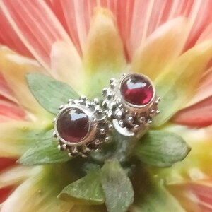 Pode incluir: Dois brincos de prata com detalhes de pedras preciosas vermelhas. Os brincos são exibidos em uma flor rosa.