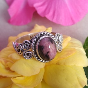 May include: A silver ring with a pink and gray oval gemstone set in a swirl design. The ring is resting on a yellow rose.