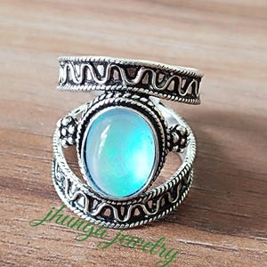 Op de afbeelding: Een zilveren ring met een ovale, iriserende, lichtblauwe edelsteen. De ring heeft een gedetailleerd, patroonontwerp rond de edelsteen en op de band. De ring ligt op een houten oppervlak. De tekst "jhungajewelry" is zichtbaar.