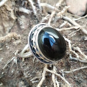 Puede incluir: Un anillo de plata con una piedra preciosa negra grande y ovalada. El anillo tiene una banda de plata detallada y un engaste decorativo alrededor de la piedra. El anillo se muestra sobre una superficie terrosa y texturizada con ramitas y pequeñas rocas.