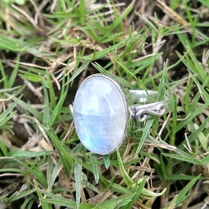 Op de afbeelding: Een zilveren ring met een grote ovale maansteen in het midden. De ring ligt op een bed van groen gras.