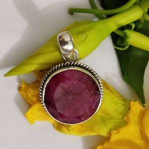 Op de afbeelding: Een zilveren hanger met een grote, gefacetteerde, dieprode edelsteen. De edelsteen is gezet in een zilveren rand met een gedraaid touwontwerp. De hanger is omgeven door gele bloemen en groene bladeren.
