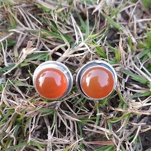 Op de afbeelding: Een paar zilveren oorbellen met oranje carneol stenen. De oorbellen zijn gezet in een eenvoudig, rond ontwerp.