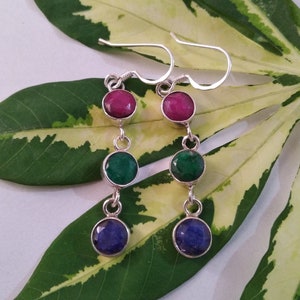 May include: Silver dangle earrings with three gemstones. The top gemstone is pink, the middle gemstone is green, and the bottom gemstone is blue.