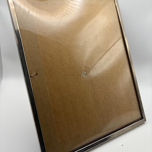 Könnte beinhalten: Ein rechteckiger Bilderrahmen mit silbernem Metallrand. Der Rahmen hat eine Holzrückwand und eine transparente Glas- oder Kunststofffront. Der Rahmen ist leer und bereit, ein Foto oder Kunstwerk anzuzeigen.
