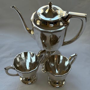 May include: A silver tea set with a teapot, a sugar bowl, and a creamer. The teapot has a lid with a decorative finial and a curved spout. The sugar bowl and creamer have handles and sit on small bases. The set is made of shiny metal.
