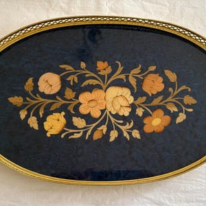 May include: An oval serving tray with a dark blue, textured surface featuring a central floral inlay design in shades of tan and gold. The tray is framed by a gold-toned metal border with a lattice pattern and includes two matching side handles.