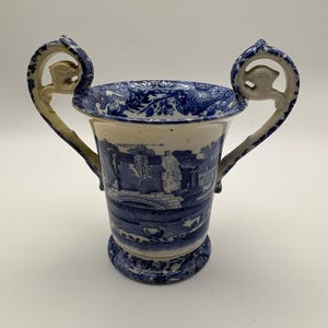 Könnte beinhalten: Eine blau-weiße Keramikvase mit zwei Henkeln. Die Vase zeigt eine Landschaftsszene und florale Muster. Die Henkel sind gebogen und haben ein dekoratives Element oben. Die Vase ist etwa 15 cm hoch.