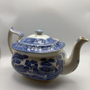 May include: A white ceramic teapot featuring a blue transferware pattern depicting a landscape with trees, a bridge, and buildings. The lid and handle are decorated with a matching blue floral motif, and the spout is curved and plain white.