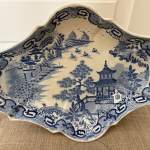 May include: A blue and white ceramic serving platter with a scalloped edge. The platter features a detailed landscape scene with pagodas, trees, and boats. The border has a repeating geometric pattern. The platter is approximately 14 inches long.