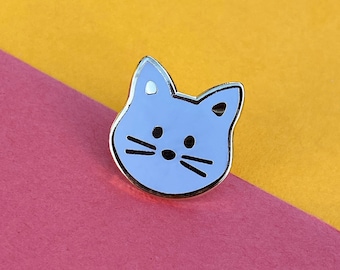 May include: A lavender blue enamel pin featuring a cartoon cat face with black eyes and whiskers.