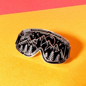 May include: A black and silver enamel pin shaped like a pair of ski goggles with a mountain scene inside. The scene includes a moon, stars, and trees.