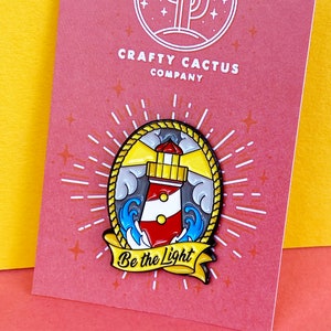 May include: A red and white lighthouse enamel pin with a yellow beam of light, surrounded by blue waves and a rope border. The pin has a yellow banner with the text "Be the Light". The pin is on a pink card with a white cactus logo and the text "Crafty Cactus Company".