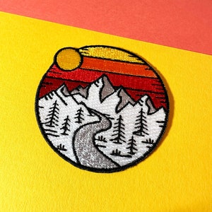 Puede incluir: Parche bordado con una escena de montaña, que presenta un río sinuoso, pinos y un sol poniente con rayas naranjas y rojas. El parche es circular y tiene un fondo blanco.
