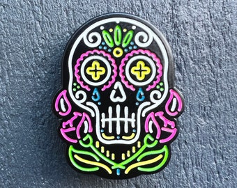 May include: Black enamel pin with a colorful sugar skull design. The skull has pink and yellow eyes, white teeth, and a floral pattern around the head. The skull is surrounded by pink and green flowers.