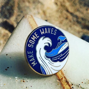 Op de afbeelding: Een ronde, marineblauwe emaille pin met de witte tekst "MAKE SOME WAVES". De pin heeft een gestileerd golfontwerp in wit en blauw. De pin heeft een goudkleurige rand en is geplaatst tegen een neutrale achtergrond.