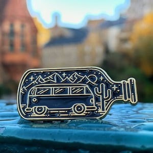 May include: A gold-coloured enamel pin shaped like a glass bottle with a black silhouette of a vintage van inside. The van is driving through a mountain range with a cactus and stars in the background.