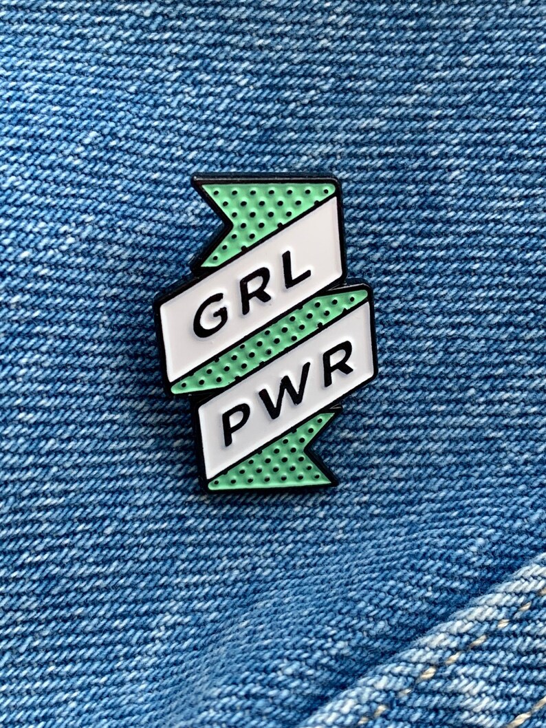 May include: Green and white enamel pin with a banner that reads "GRL PWR" in white letters. The banner has a dotted pattern.