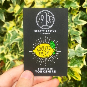 May include: A black and white enamel pin featuring a yellow lemon with green leaves and the text "Squeeze the Day". The pin is designed in Yorkshire and is sold by Crafty Cactus Company.