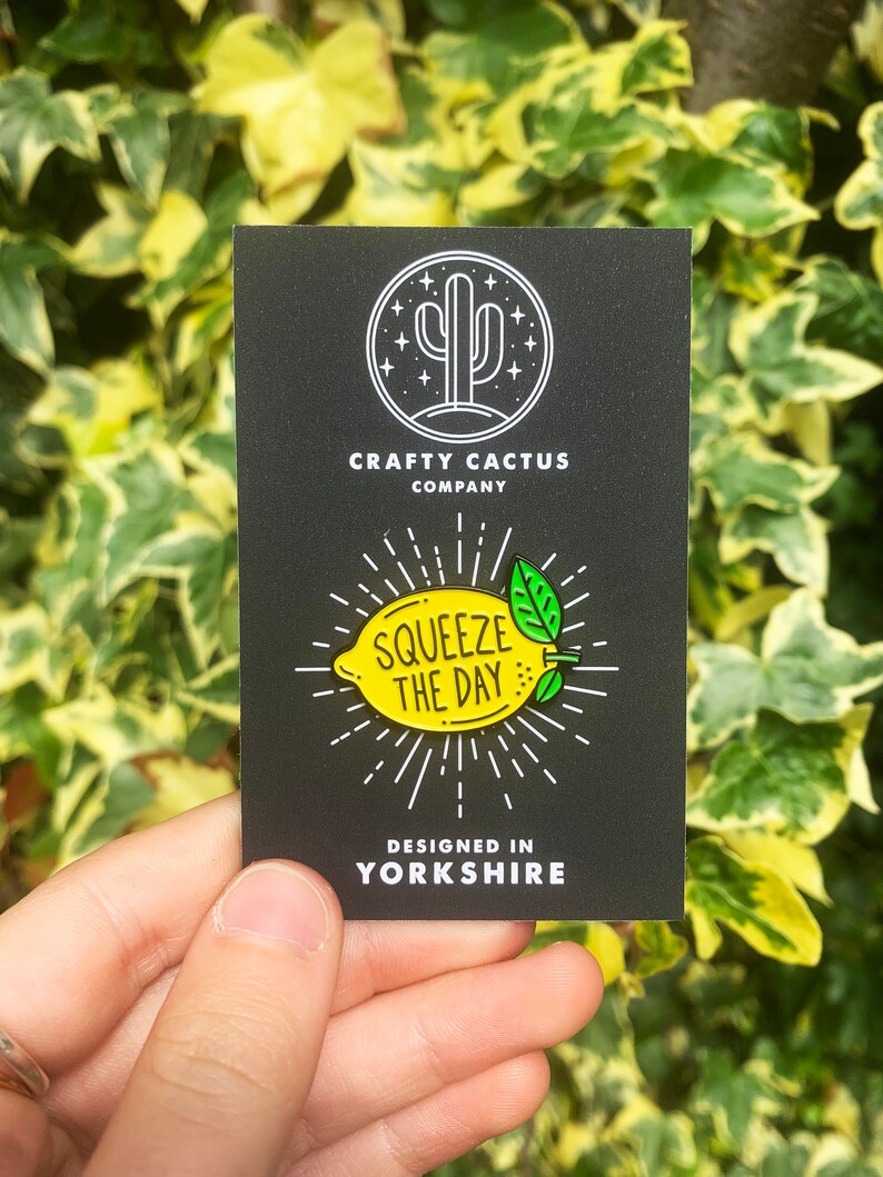 May include: A black and white enamel pin featuring a yellow lemon with green leaves and the text "Squeeze the Day". The pin is designed in Yorkshire and is sold by Crafty Cactus Company.