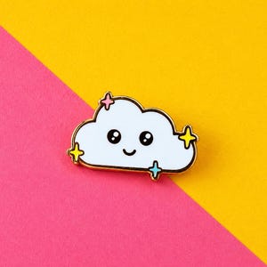 May include: A white enamel pin shaped like a smiling cloud with black eyes and a small mouth. The cloud features four small, colourful four-pointed star accents in pink, yellow, and light blue, all outlined in a thin gold-toned metal border.