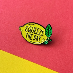 May include: Yellow enamel pin shaped like a lemon with a green leaf. The lemon has the text "Squeeze the Day" written on it.