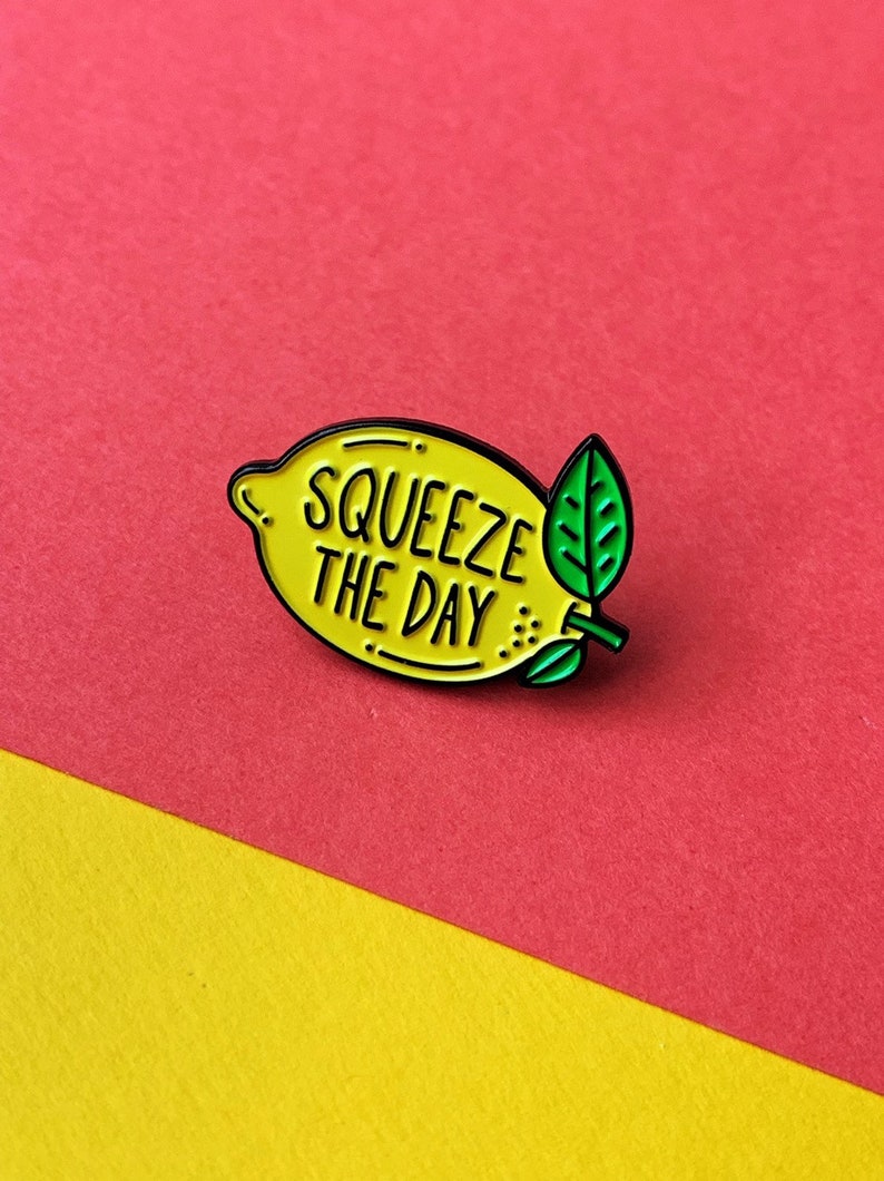 May include: Yellow enamel pin shaped like a lemon with a green leaf. The lemon has the text "Squeeze the Day" written on it.