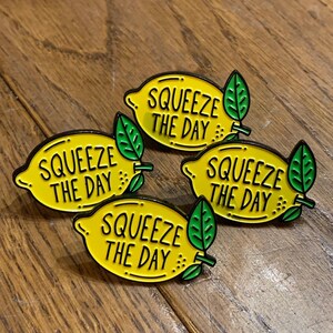 May include: Four yellow lemon-shaped enamel pins with black outlines and green leaves. Each pin says "Squeeze the Day" in black lettering.